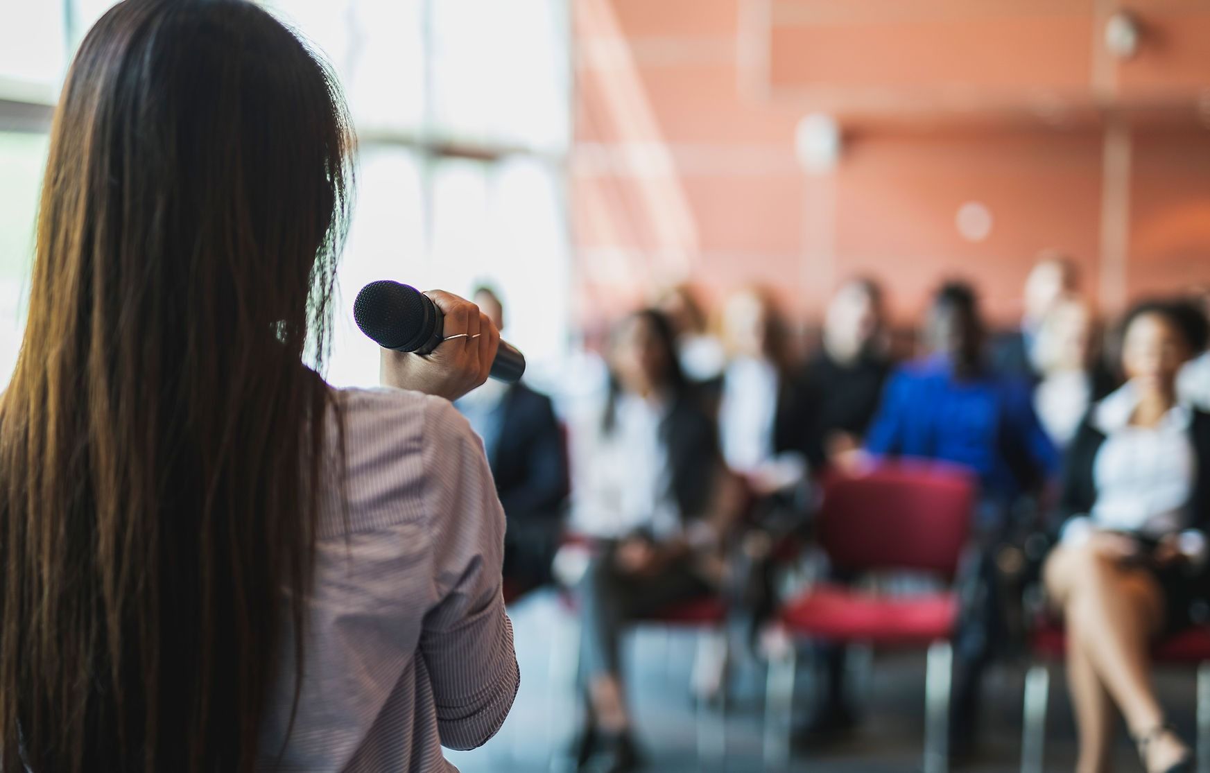 A speaker stands before an audience in a conference room, holding a microphone while facing away from the camera.