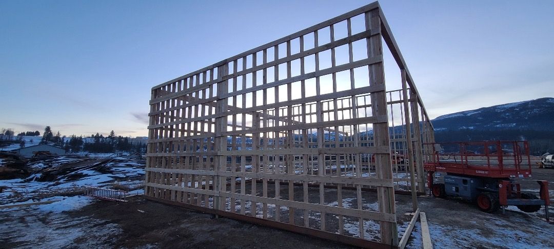 Wooden frame of a building under construction on a snowy site, with a lift truck and mountain backdrop.
