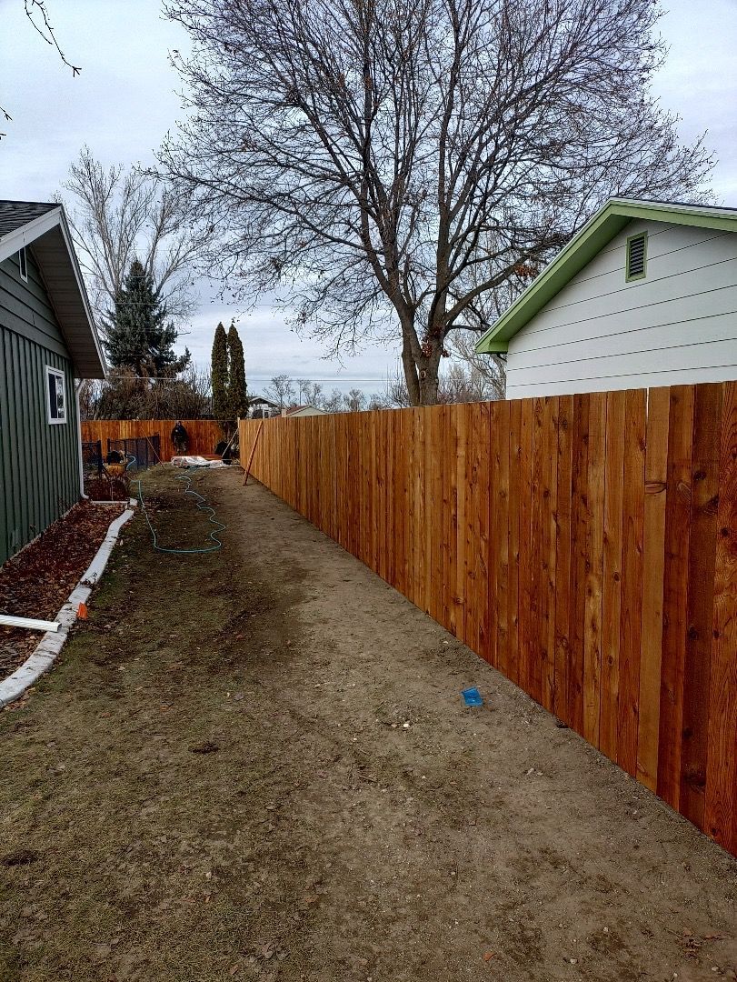 Wooden fence bordering a yard with brown grass and small buildings, under a cloudy sky.