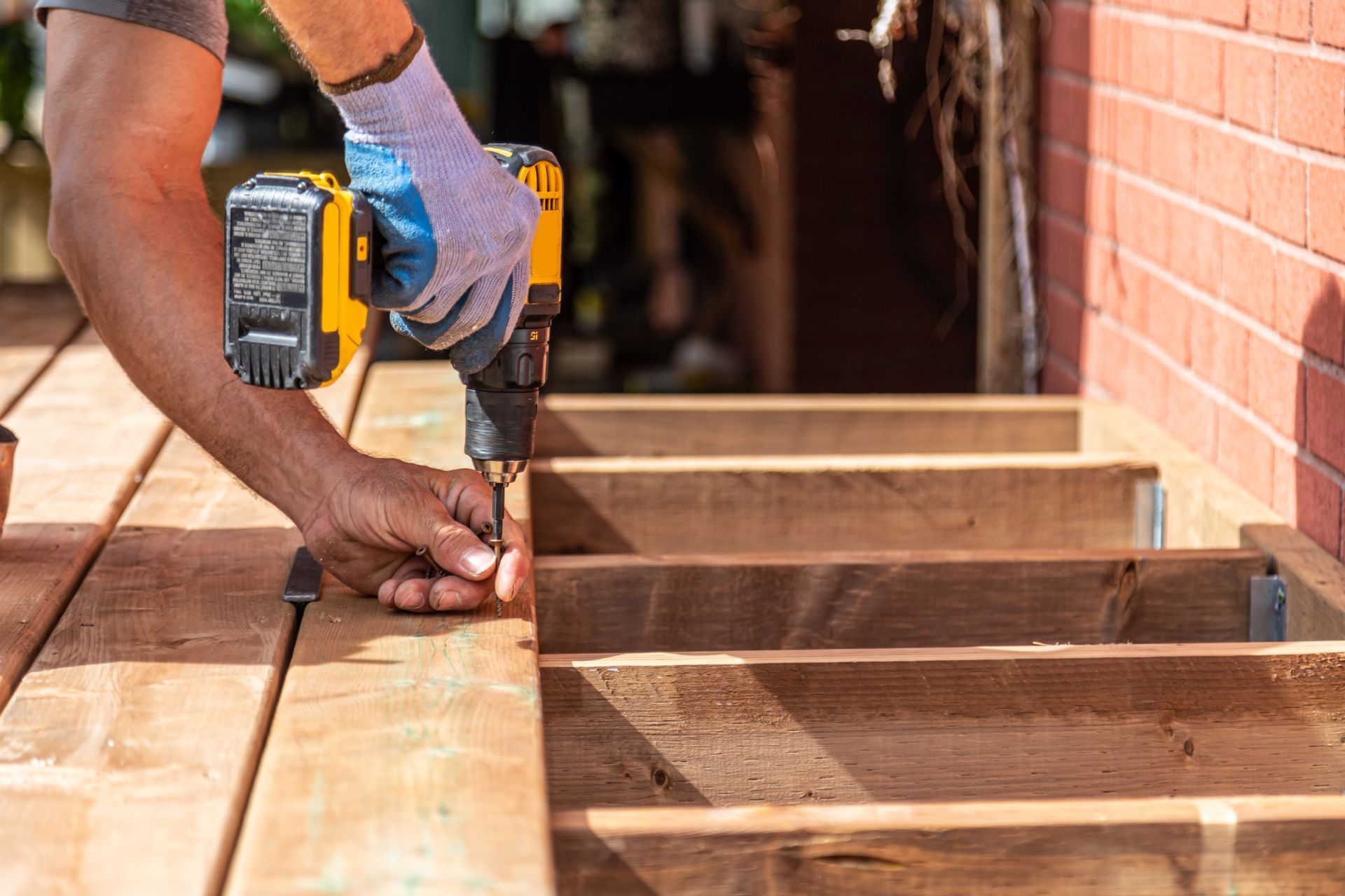 A person in a blue glove uses a yellow power drill to secure a wooden board onto a deck frame next to a brick wall.