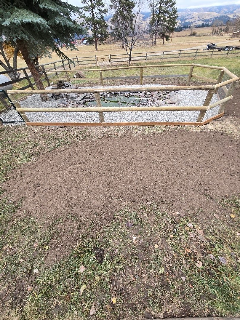 A fire pit enclosed by a wooden fence, on gravel, with dirt area in front, and a grassy field background.