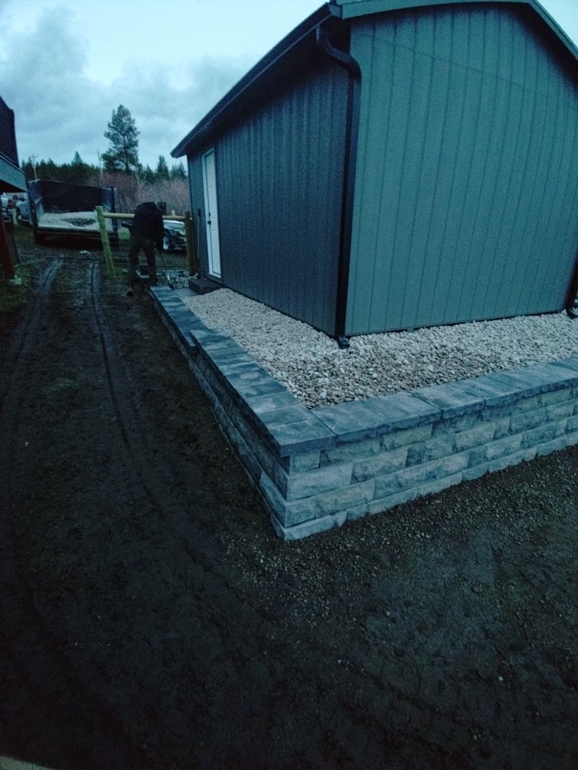 A shed with a gray retaining wall and gravel. A person is near a wooden fence.