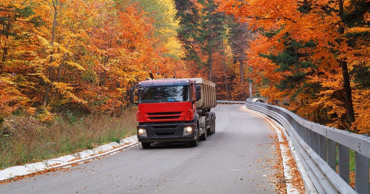 A red dump truck is driving through a curved road, cutting through a forest with orange and yellow trees.