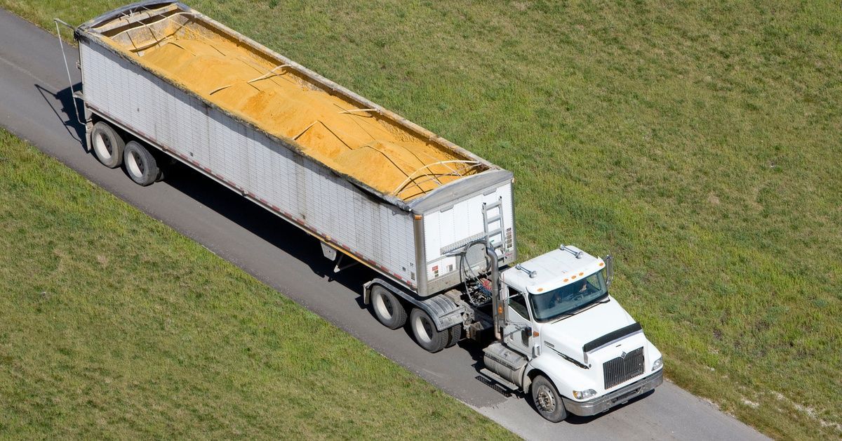 An aerial shot of a fleet truck driving down a single lane road hauling an open trailer of corn.