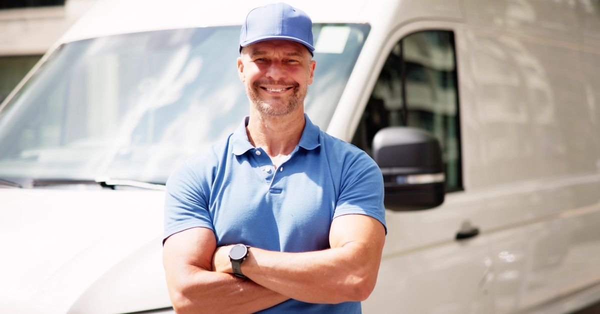 A man wearing a light blue cap and a polo while smiling. He is standing in front of a large, white van.