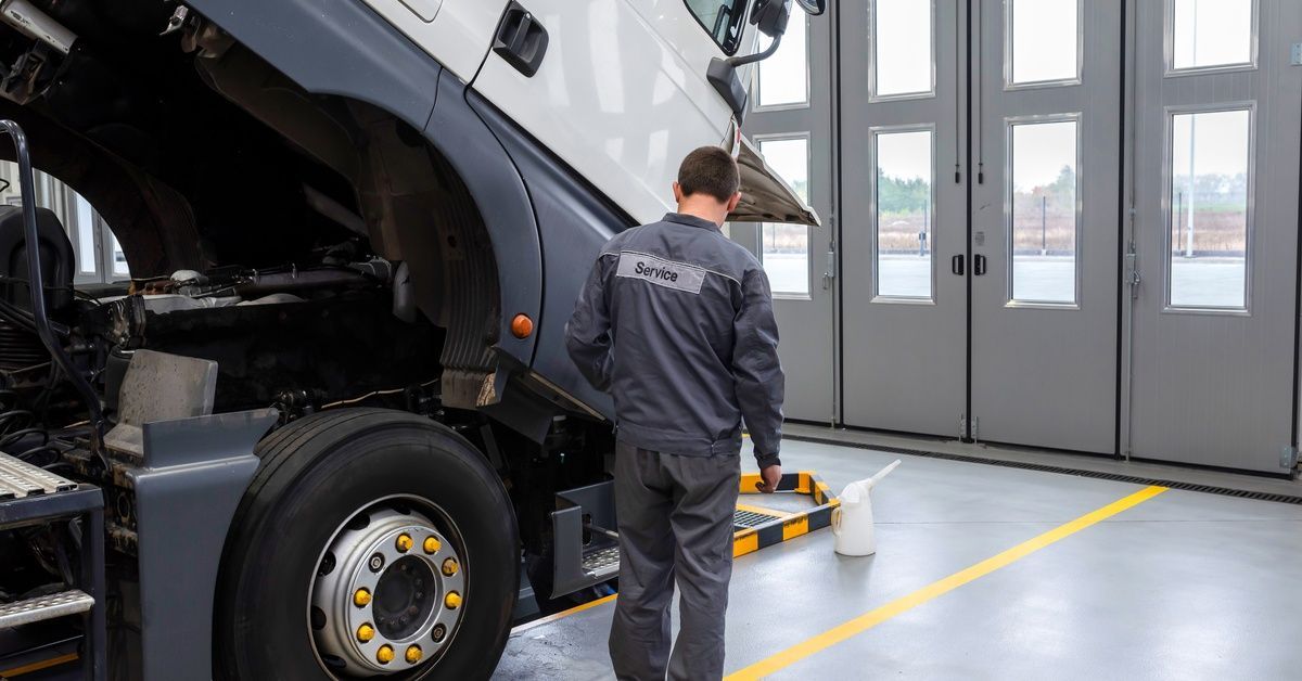 A mechanic is at work inspecting the front of a trailer truck's cabin. The cabin is being lifted by an out-of-frame machine.