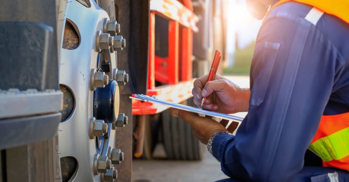 A focus shot of the wheel of a trailer truck. A worker is kneeling down, taking notes on a clipboard.