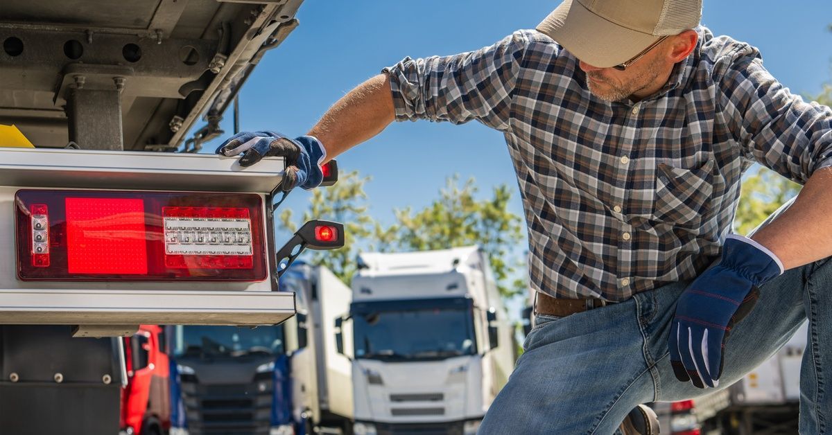 A man in denim jeans, flannel, sunglasses, a cap, and work gloves bending down to inspect a truck.