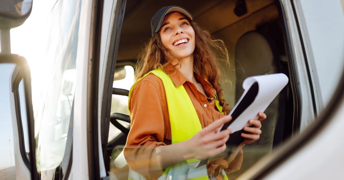 A woman wearing a cap and an orange high-visibility vest, smiling and sitting in a semi-truck driver's cabin, holding a clipboard.