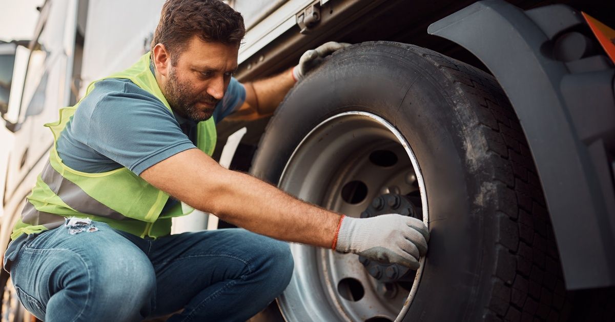 A worker in a neon green high-visibility vest and gloves, bending down to inspect a semi-truck tire.