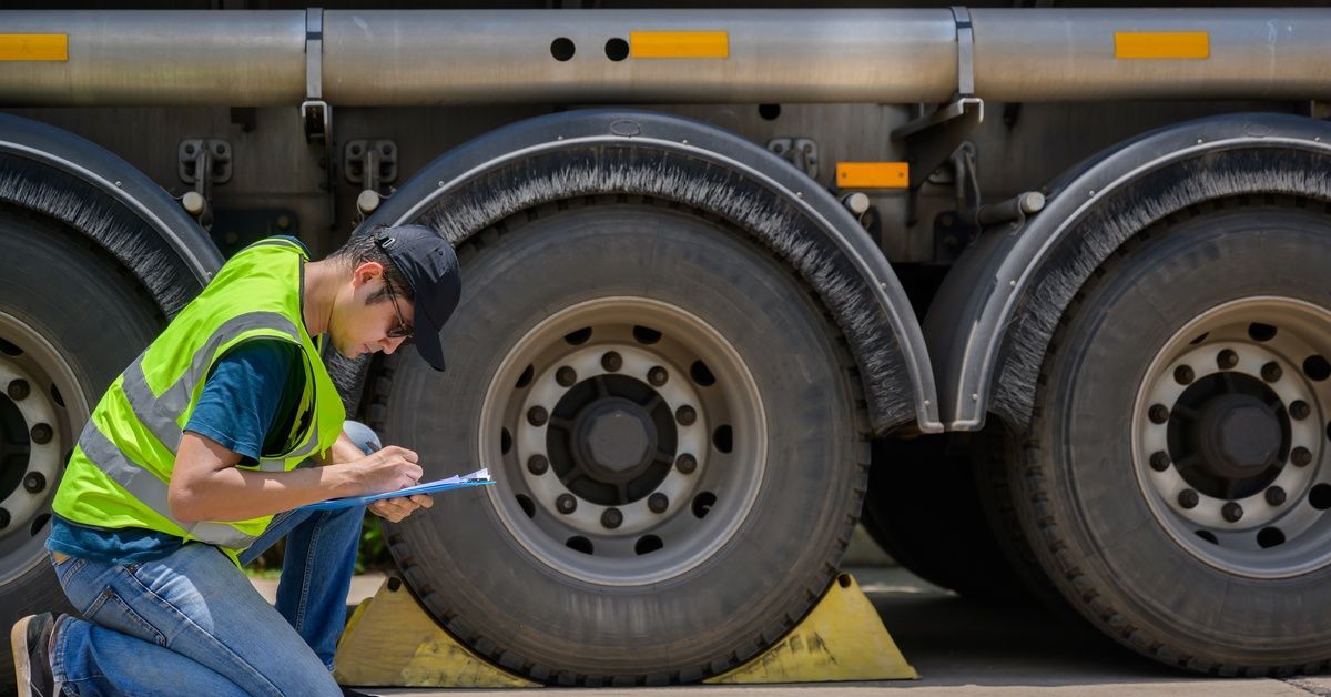 A man in a black cap and high-visibility vest, kneeling in front of semi-truck wheels as he takes notes on a clipboard.