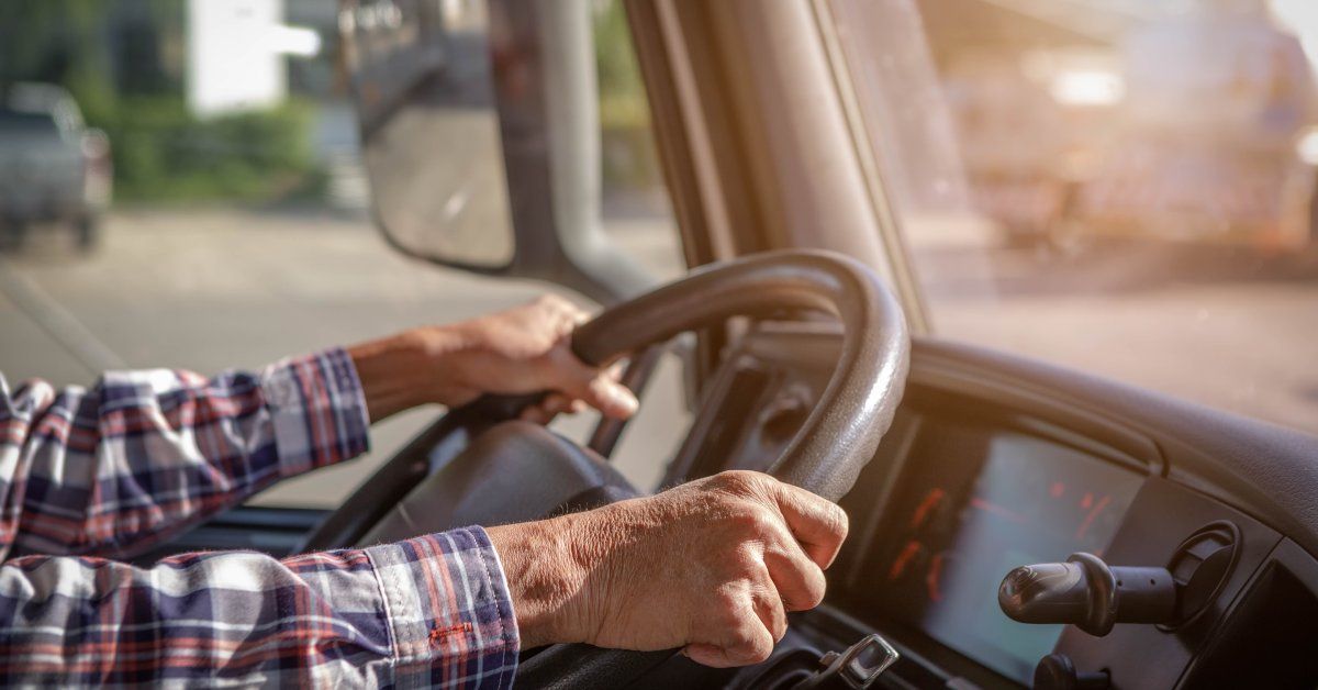 A close-up of an older man's hands holding the wheel of a semi-truck as he drives through a parking lot.