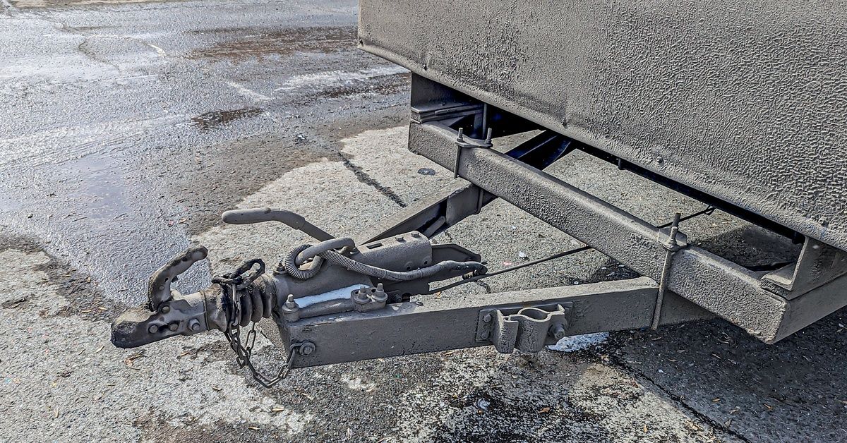 The gray metal tow bar of a cargo trailer in a gray asphalt parking lot. There is nothing attached to the tow bar of the cargo trailer.