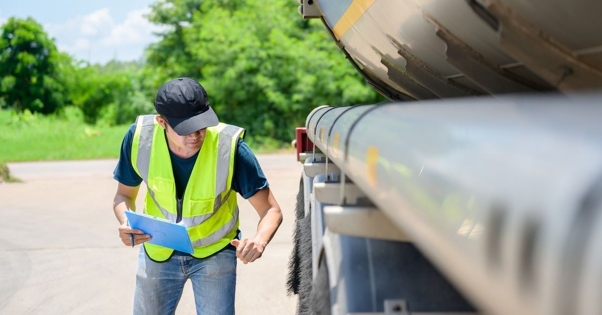 A worker wearing a cap and a green, high-visibility vest examines the wheels of a truck while holdin