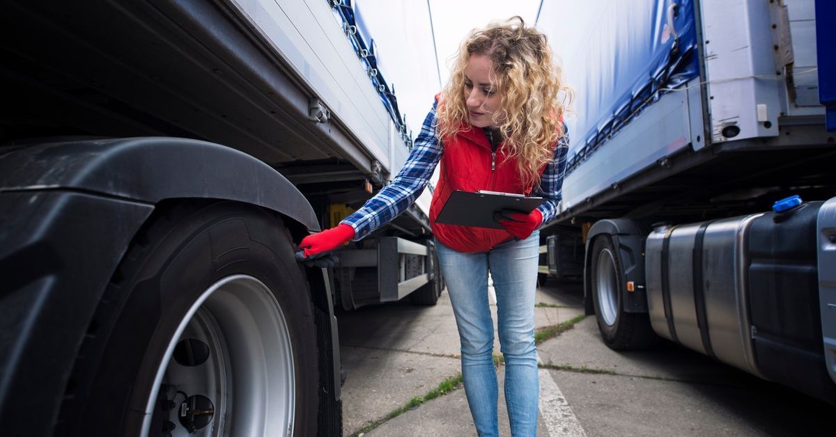 A woman wearing a flannel and a puffer vest holds a clipboard, while bending over to inspect the wheels of a truck.