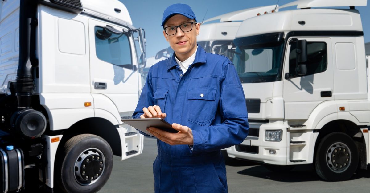 A fleet manager in blue overalls stands with a tablet in his hands in front of three parked semitrai