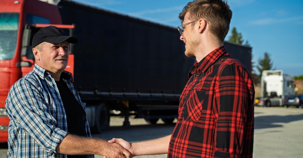 Two working men in flannels shake hands in a parking lot in front of a long red semitrailer.