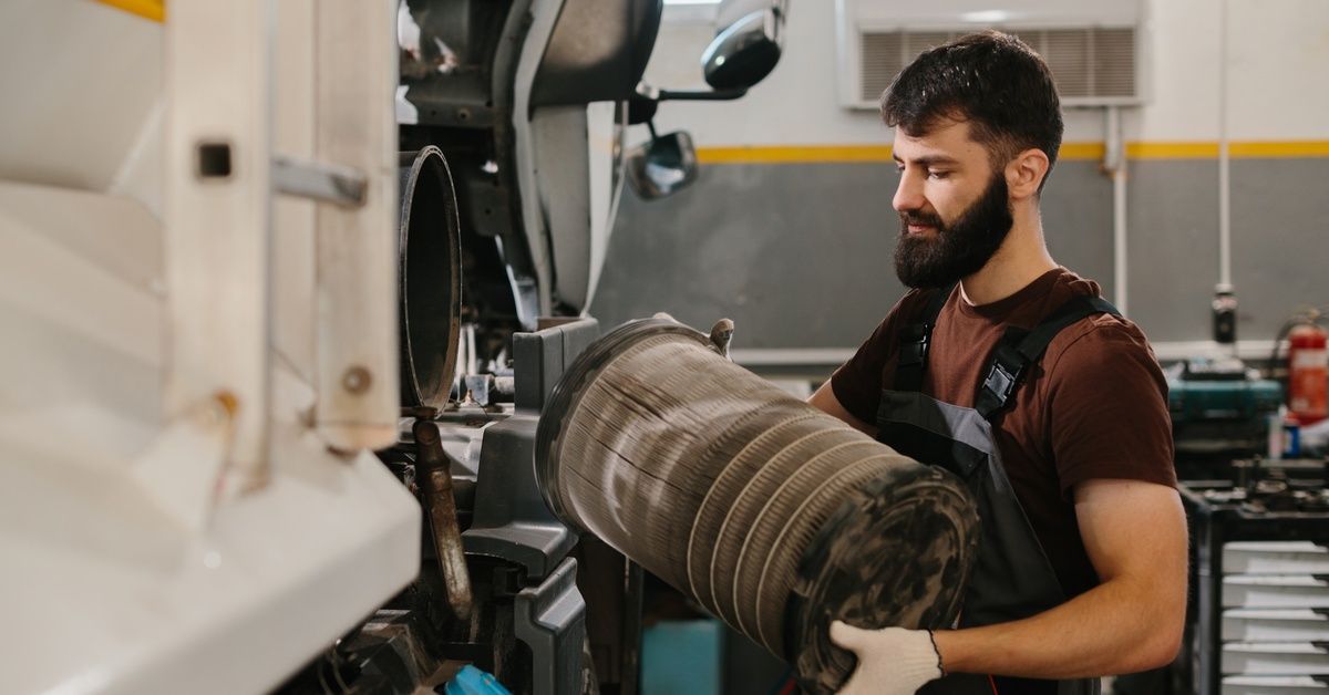 A mechanic in gray overalls and a burgundy T-shirt removes the huge air filter from the side of a semitrailer in a garage.