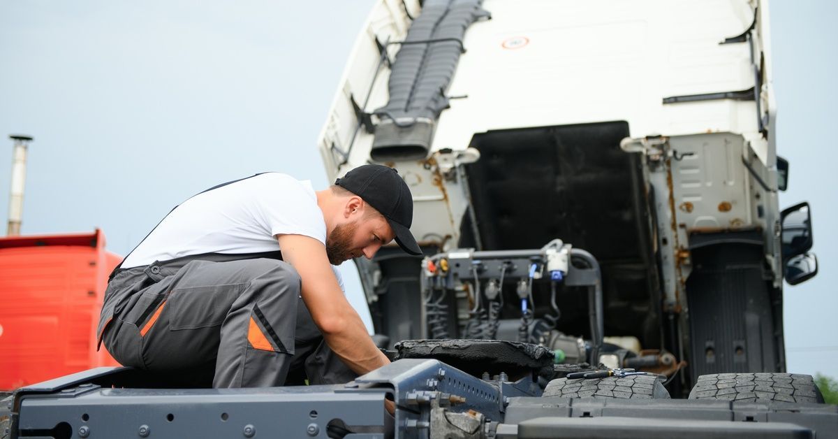 A mechanic in gray overalls and a black cap crouches near the fifth wheel housing of a white semitrailer to perform repairs.