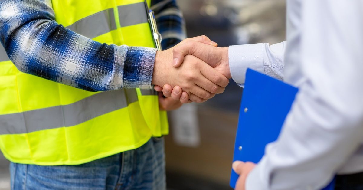 A man in jeans, flannel, and a yellow safety vest shakes the hand of a man in business attire.