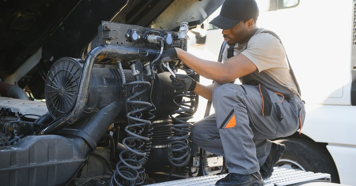 A mechanic in overalls kneeling on a semitruck's bed and using a wrench to adjust part of the air brake system.