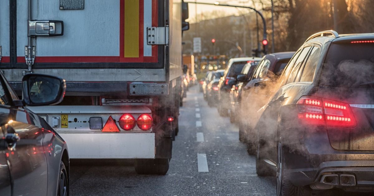 Traffic backs up behind a delivery truck emitting exhaust fumes that rise in a cold, sunset-lit lane