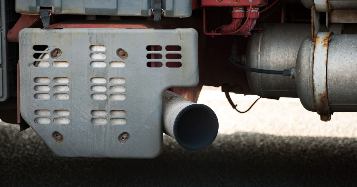 A close-up of a large truck's exhaust pipe under the chassis, beside a metal heat shield and tank.