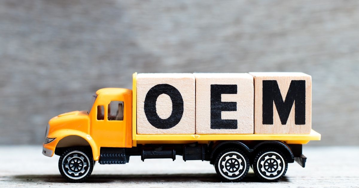 A yellow toy truck carries wooden blocks spelling OEM on its bed against a softly blurred background