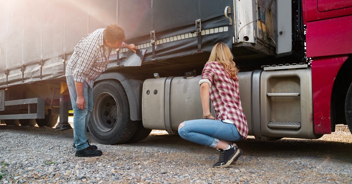 A man and woman inspect the side of a red semitruck parked on a gravel lot, checking the trailer and fuel tank.