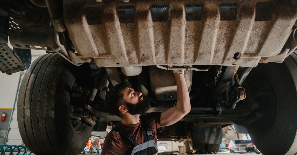 A mechanic stands beneath a raised semitruck, reaching up to inspect the undercarriage between the rear wheels.