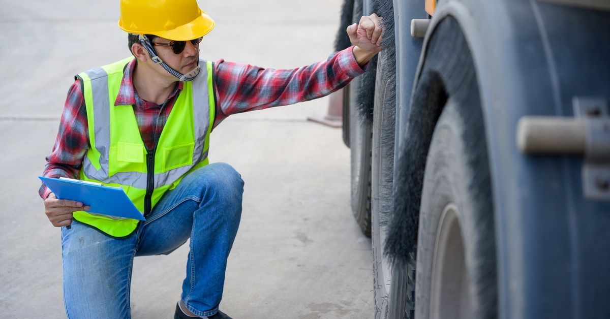 A worker in a hard hat and safety vest kneels beside a truck, inspecting the tire while holding a cl