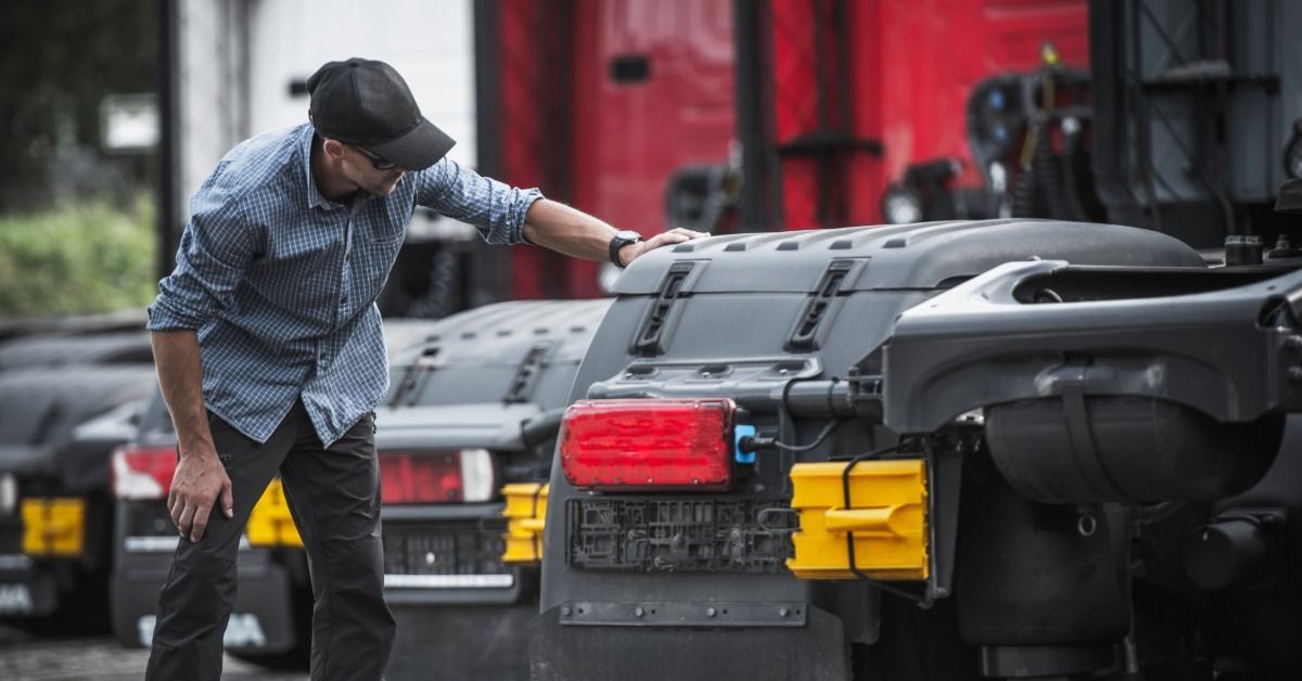 A man in a cap inspects the rear of a parked semitruck, leaning over the chassis in a full truck yard.