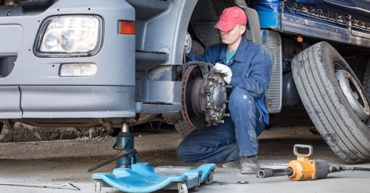 A mechanic kneels by a jacked-up blue semitruck, repairing the exposed wheel hub with tools on the ground.
