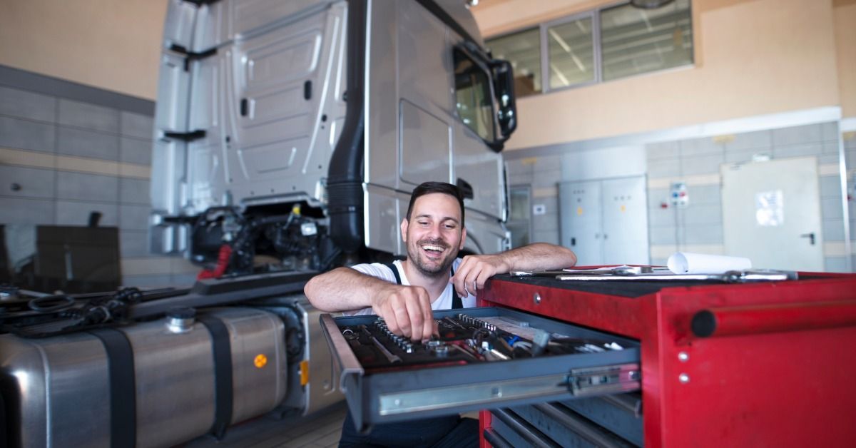 A smiling mechanic reaches into a red tool chest beside a semitruck he's servicing in a repair garage.