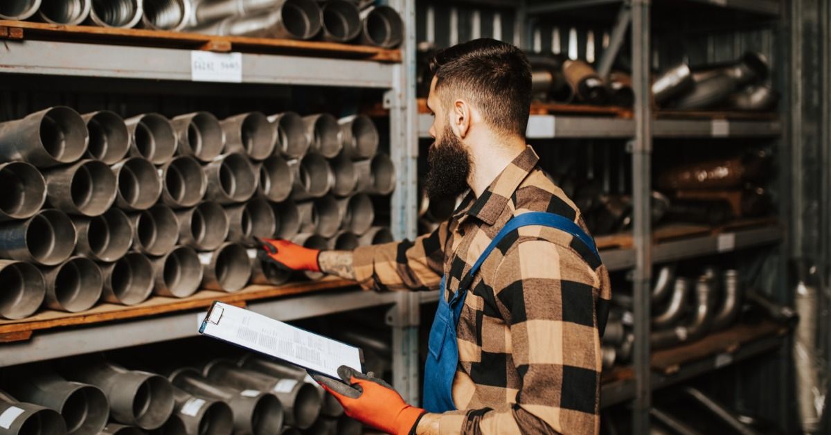 A worker holding a clipboard checks metal tube parts on warehouse shelves in a storage aisle.