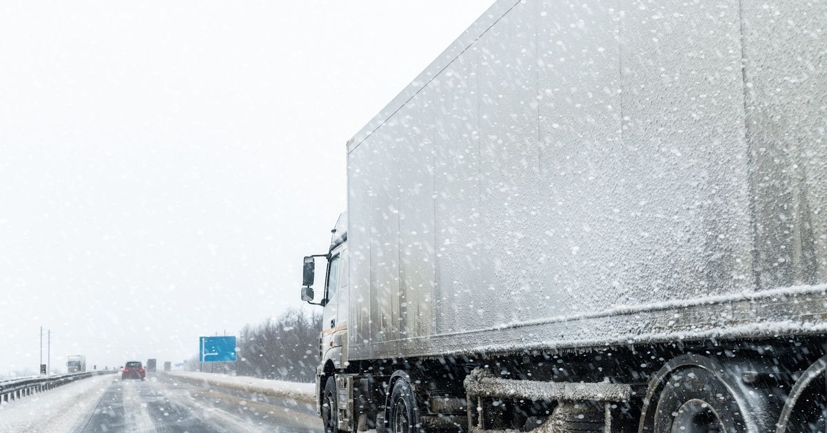 A side view of a semi-truck driving down a snowy highway. Large snowflurries hang in the air with other vehicles on the road.
