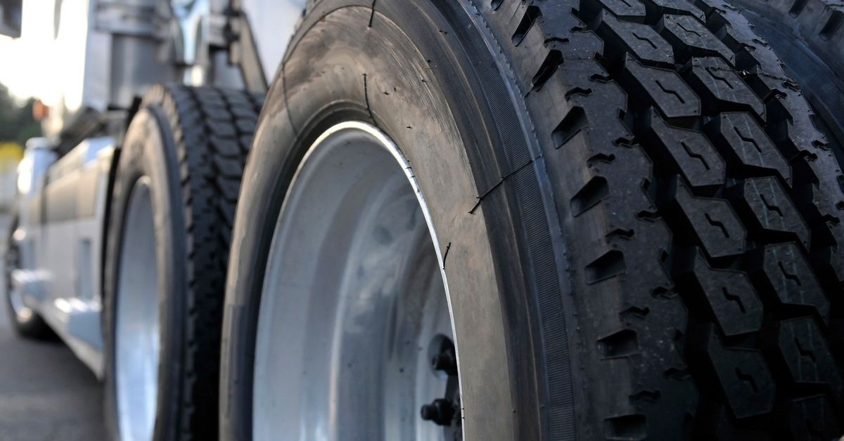 A close-up of rear semi-truck tires with deep tread and shiny metal rims. The chassis and body are visible in the background.