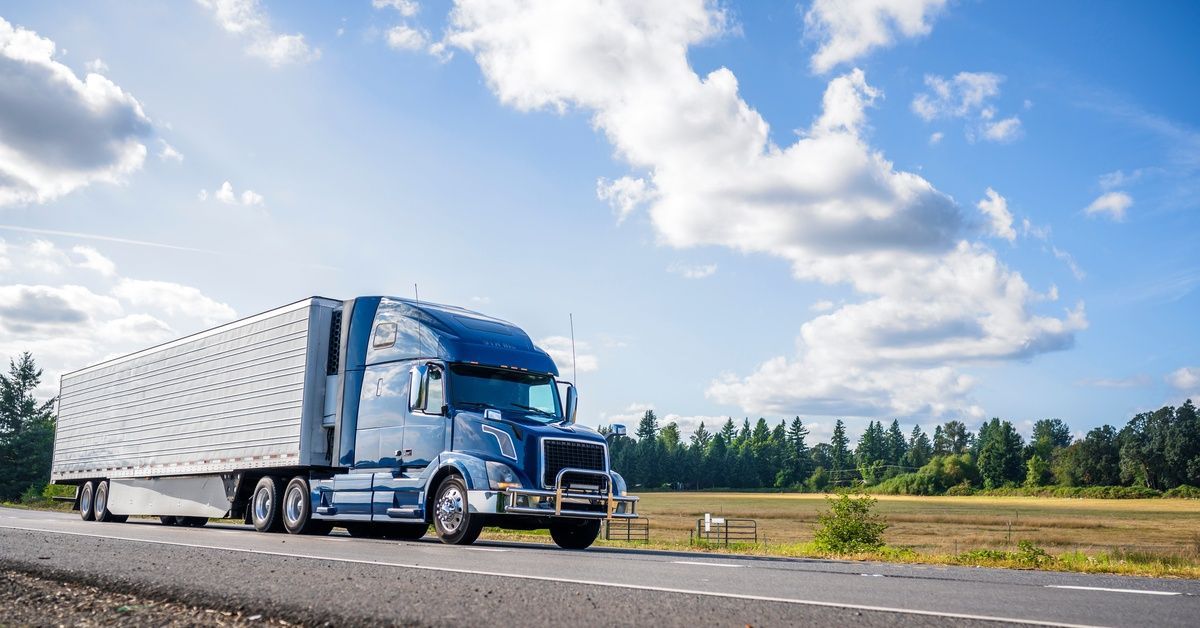 A blue semi-truck with a silver trailer. The truck drives on a rural road under a bright blue sky.