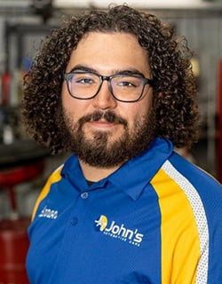 Man with curly hair and glasses wearing a blue and yellow polo shirt, standing in an auto shop.