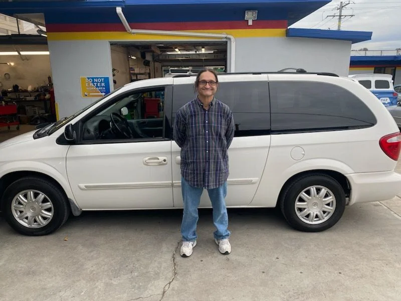 Man standing in front of a white minivan at a car repair shop; cloudy sky.