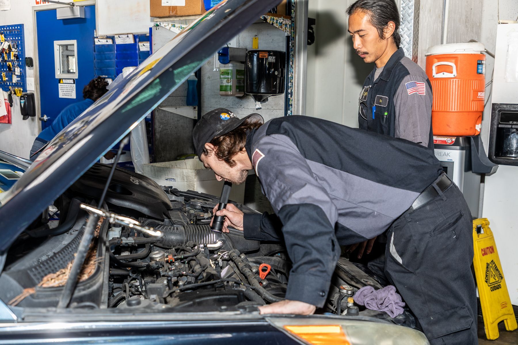 Mechanics working on a car engine in a repair shop. One leans in close, while another observes.