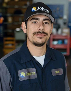 Man in John's Auto Service uniform and cap smiles. Inside a shop setting.