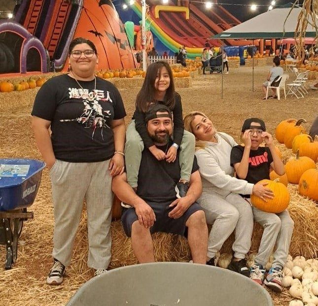 Family at a pumpkin patch. Adults and kids, some with pumpkins, near hay bales and inflatables.