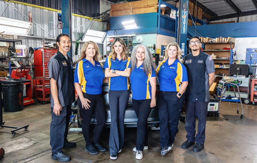 Auto repair shop team posing: six people in blue/yellow shirts, smiling, standing in front of a car lift.