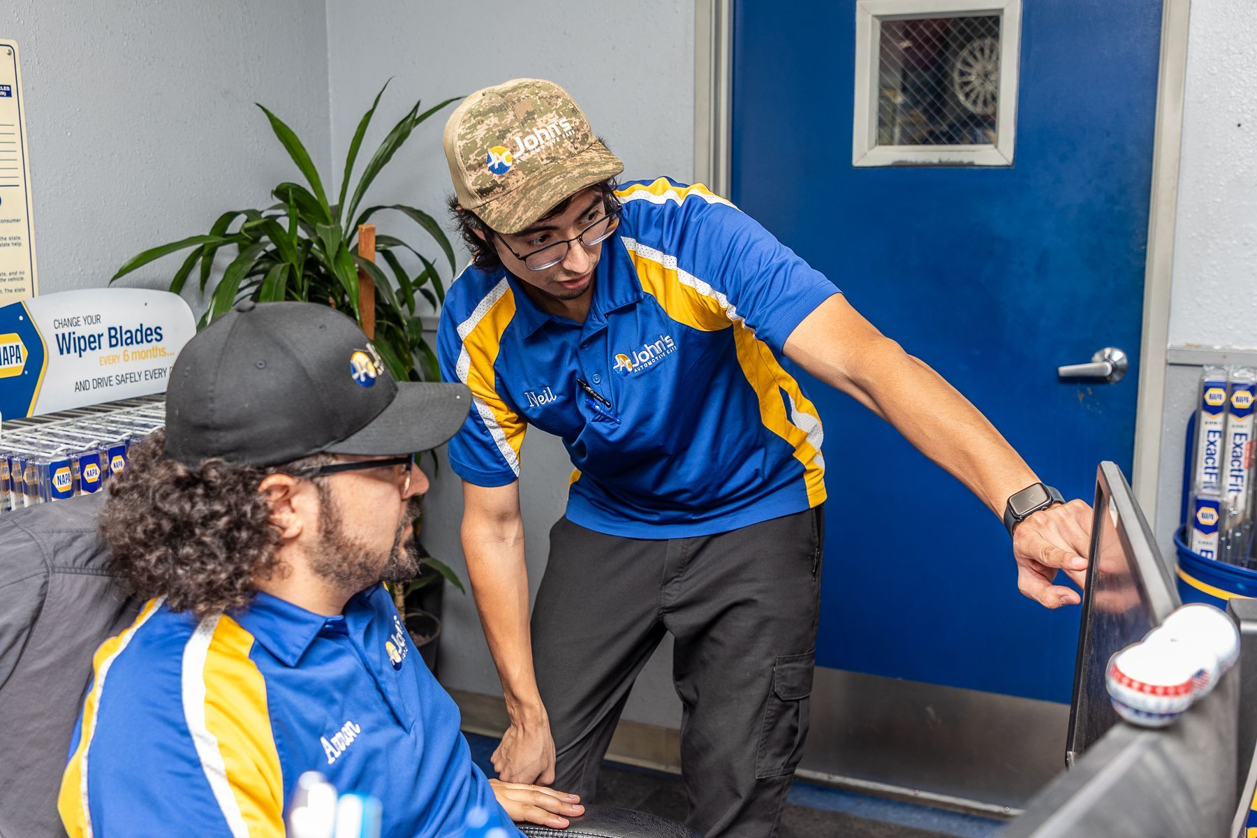 Two men in blue & yellow work shirts at a computer, one pointing, indoors.