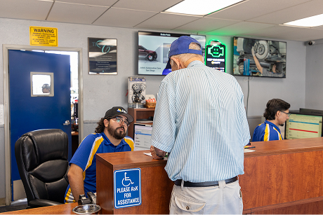 Man at auto repair shop counter talking to a worker. Other worker at a computer, shop interior.