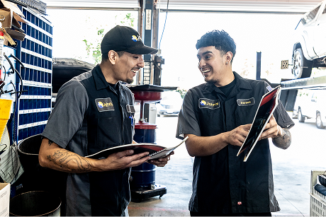 Man at auto repair shop counter talking to a worker. Other worker at a computer, shop interior.