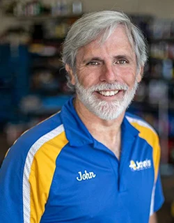 Man with gray hair and beard wearing a blue shirt with yellow trim, smiling in a shop setting.