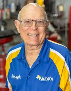 Smiling man wearing glasses and a blue and yellow mechanic shirt, in a garage.