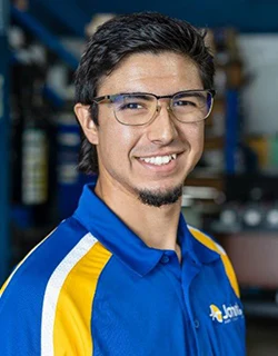 Man with glasses smiles, wearing blue and yellow collared shirt in a workshop setting.